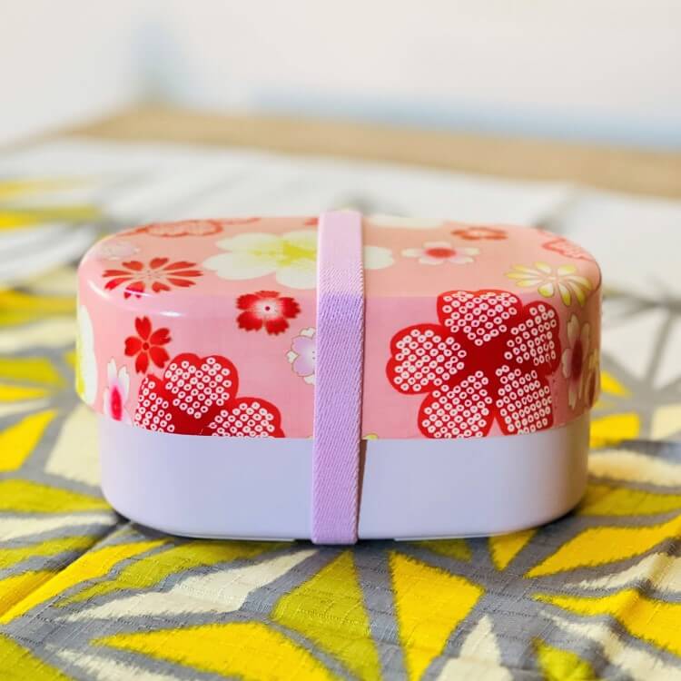 side photo of pink bento box with sakura and flower patterns secured with pink lunch band