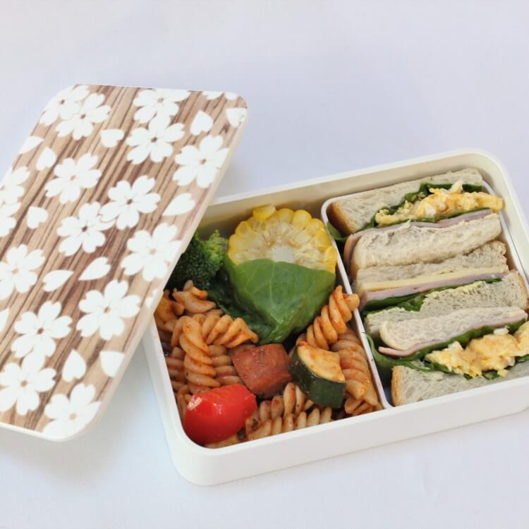 Overhead shot of white sakura mokume bento box with sakura flowers on the lid and showing pasta and sandwich lunch